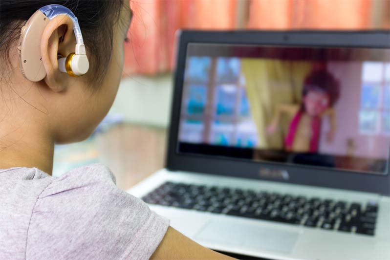 Looking over the shoulder of a young girl who is wearing a large hearing aid while she watches cartoons on her laptop.