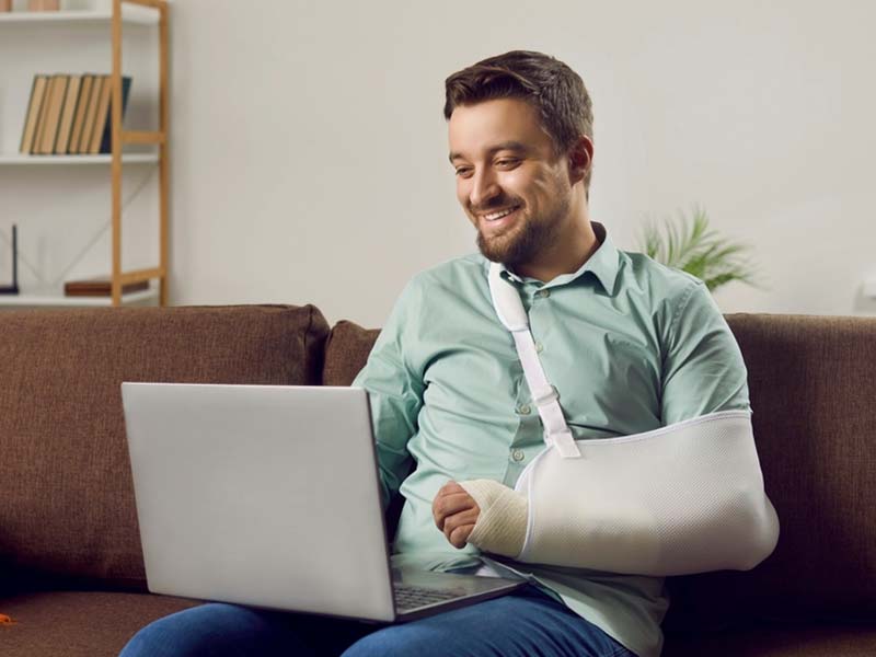 Happy man with his arm in a sling and cast on the couch using a laptop