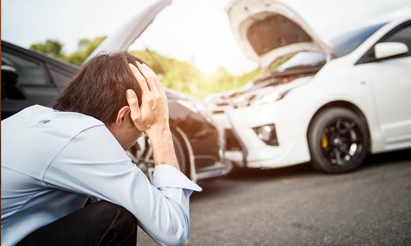 Man crouched down holding his head with smashed cars in the background