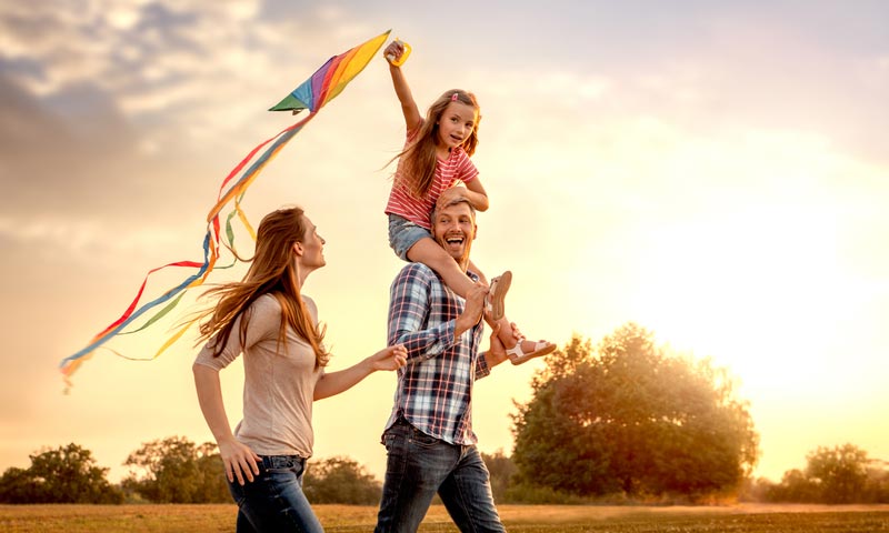 Young parents with daughter on fathers shoulders flying kite at sunset