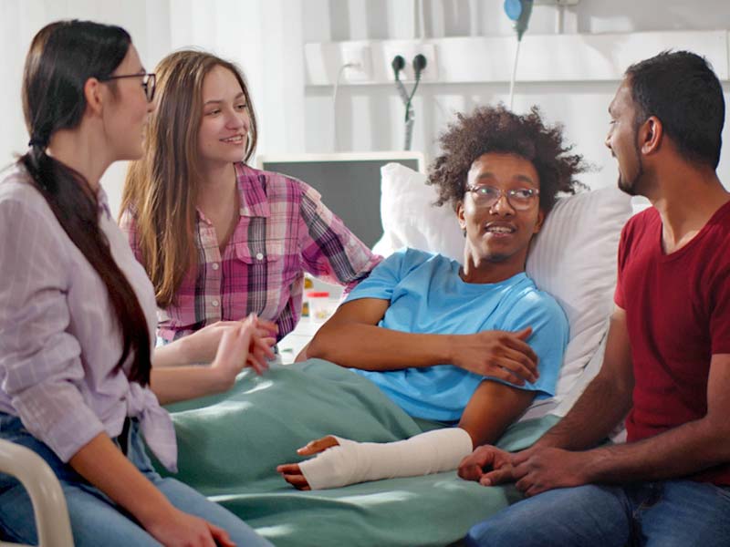 A young man laying in a hospital bed with three friends hanging out bedside