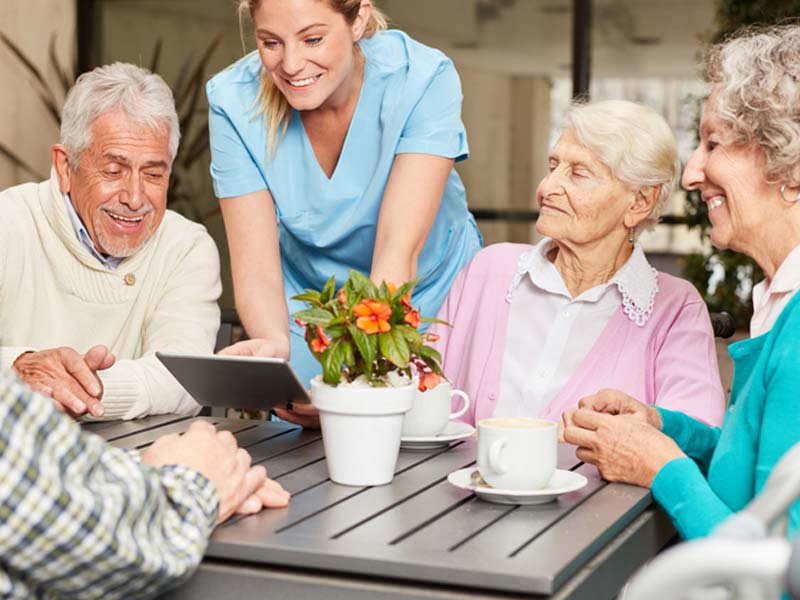 Four seniors, men and women around a table playing with a nurse's tablet